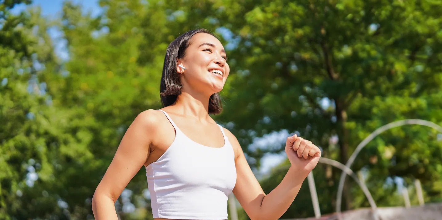 A woman in a white tank top smiles while exercising outdoors on a sunny day, with green trees in the background.