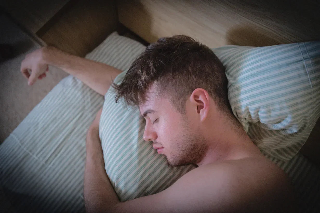 A young man is sleeping on his side in bed, resting his head on a striped pillow with his arm draped over the bed.