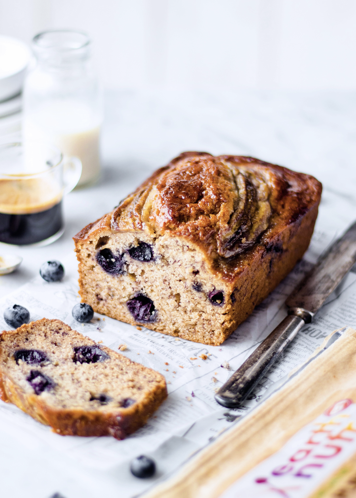 Blueberry loaf cake with a marbled top on parchment paper, with a sliced piece in front, fresh blueberries scattered nearby, and a coffee and milk in the background.