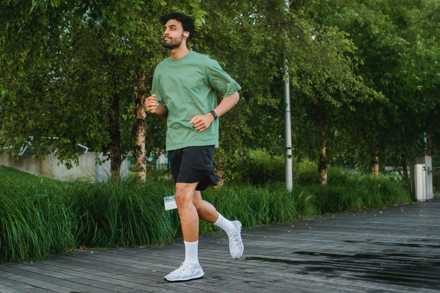 A man jogs outdoors on a wooden pathway surrounded by tall green grass and leafy trees, wearing a green T-shirt, black shorts, white socks, and white running shoes, with wireless earbuds and a fitness watch.