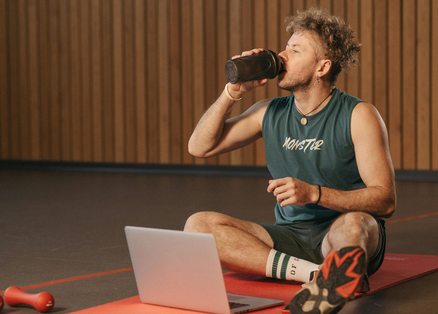Person sitting on a red exercise mat in a gym, drinking from a black shaker bottle after a workout. A laptop and small dumbbell rest nearby on the floor, suggesting a virtual workout session.