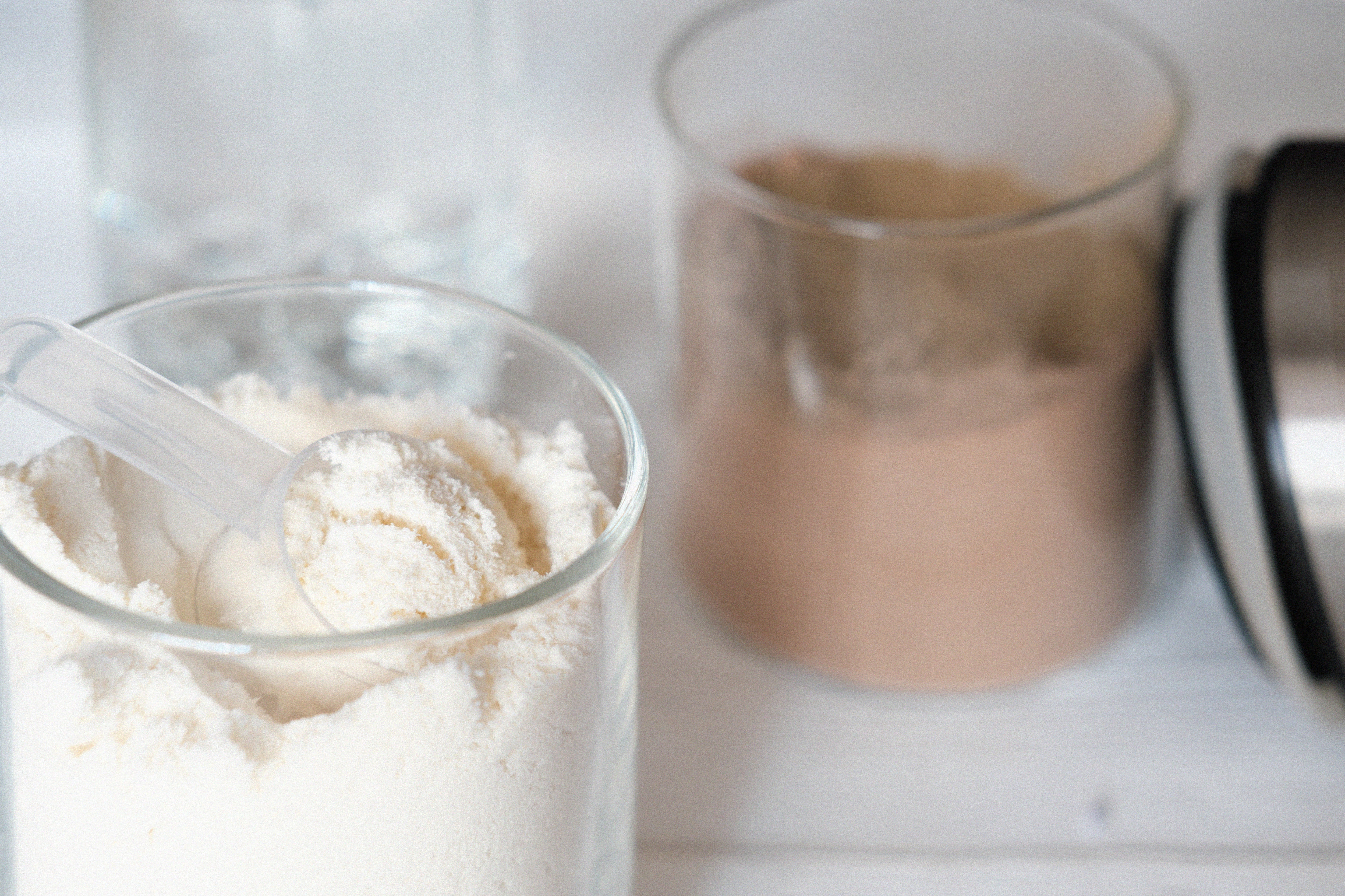 Clear glass filled with white powder and a transparent scoop in the foreground, with a second glass containing a mixed light-brown beverage softly blurred in the background on a white surface