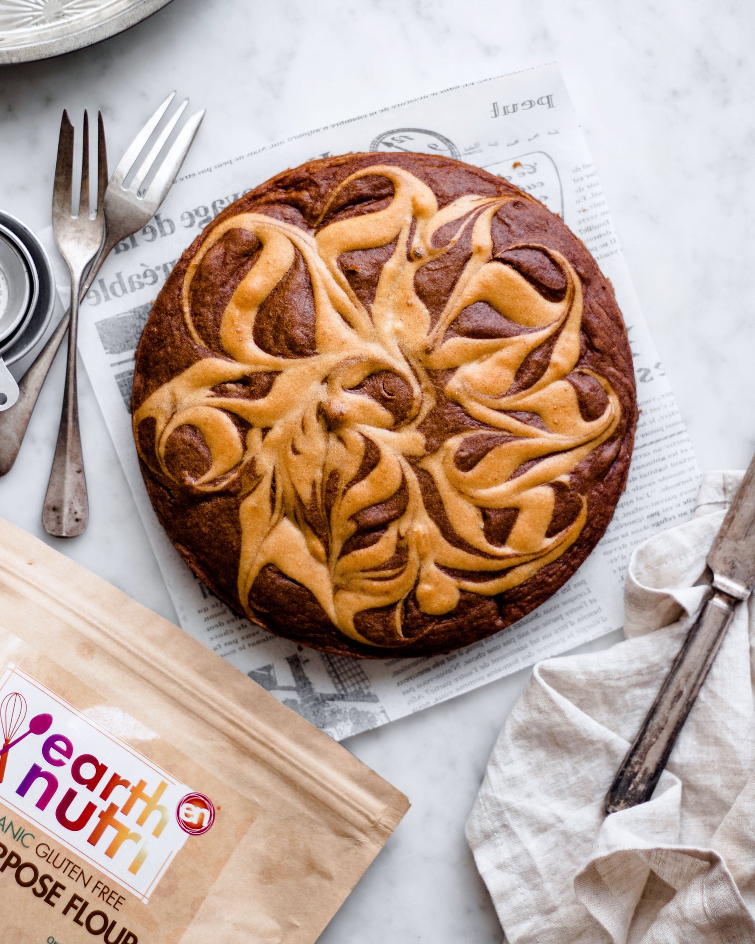 Swirled cinnamon-style cake on parchment paper, topped with a decorative glaze pattern, with utensils and an EarthNutri multipurpose flour bag on a light surface.