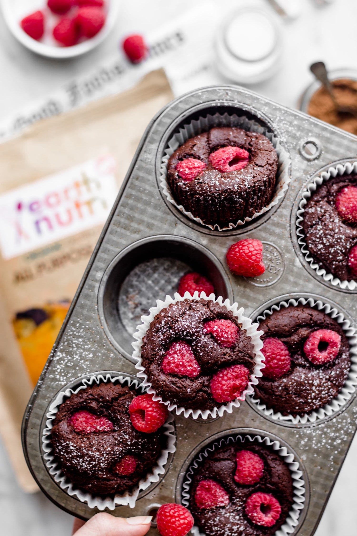 Chocolate muffins topped with fresh raspberries in a metal muffin tin, with extra raspberries and ingredients scattered on a light surface.