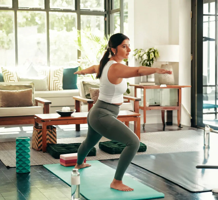 alt text. Woman practicing a yoga lunge pose on a mat in a bright living room, with foam rollers, blocks, and a water bottle nearby.