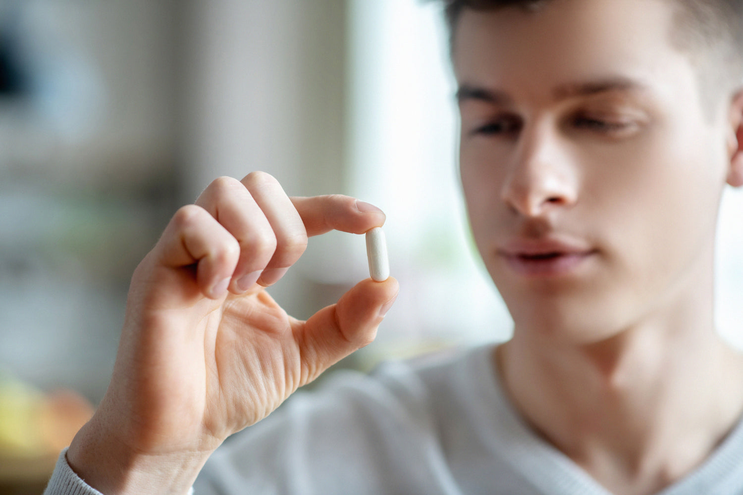 A young man holds a white capsule between his thumb and index finger, examining it closely with a soft, blurred background behind him.