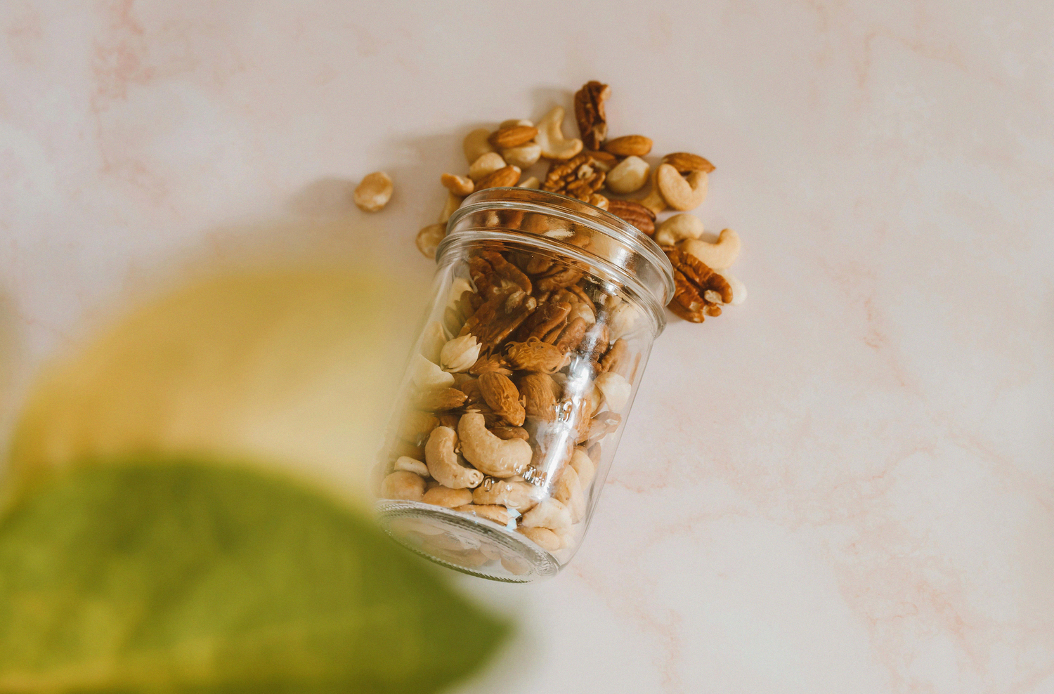 Glass jar tipped on its side with mixed nuts spilling out onto a light marble surface, with a soft, blurred green leaf in the foreground.