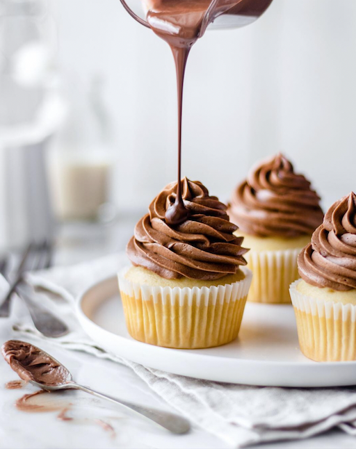 Vanilla cupcakes topped with swirled chocolate frosting as melted chocolate is poured over one, with a spoon and soft neutral background.