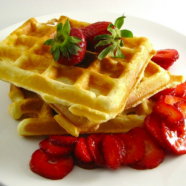 Stack of golden waffles topped with whole strawberries and served with sliced strawberries on a white plate.