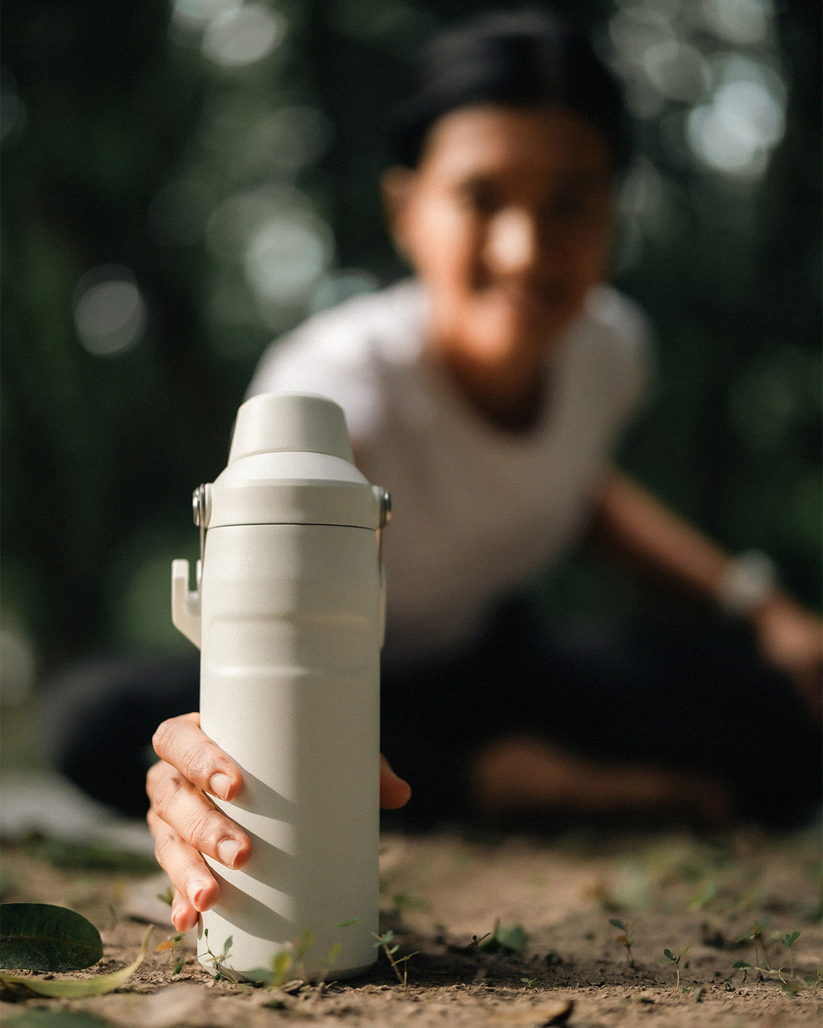 Close-up of a reusable white bottle being reached for outdoors, with a blurred person in the background in a natural setting.