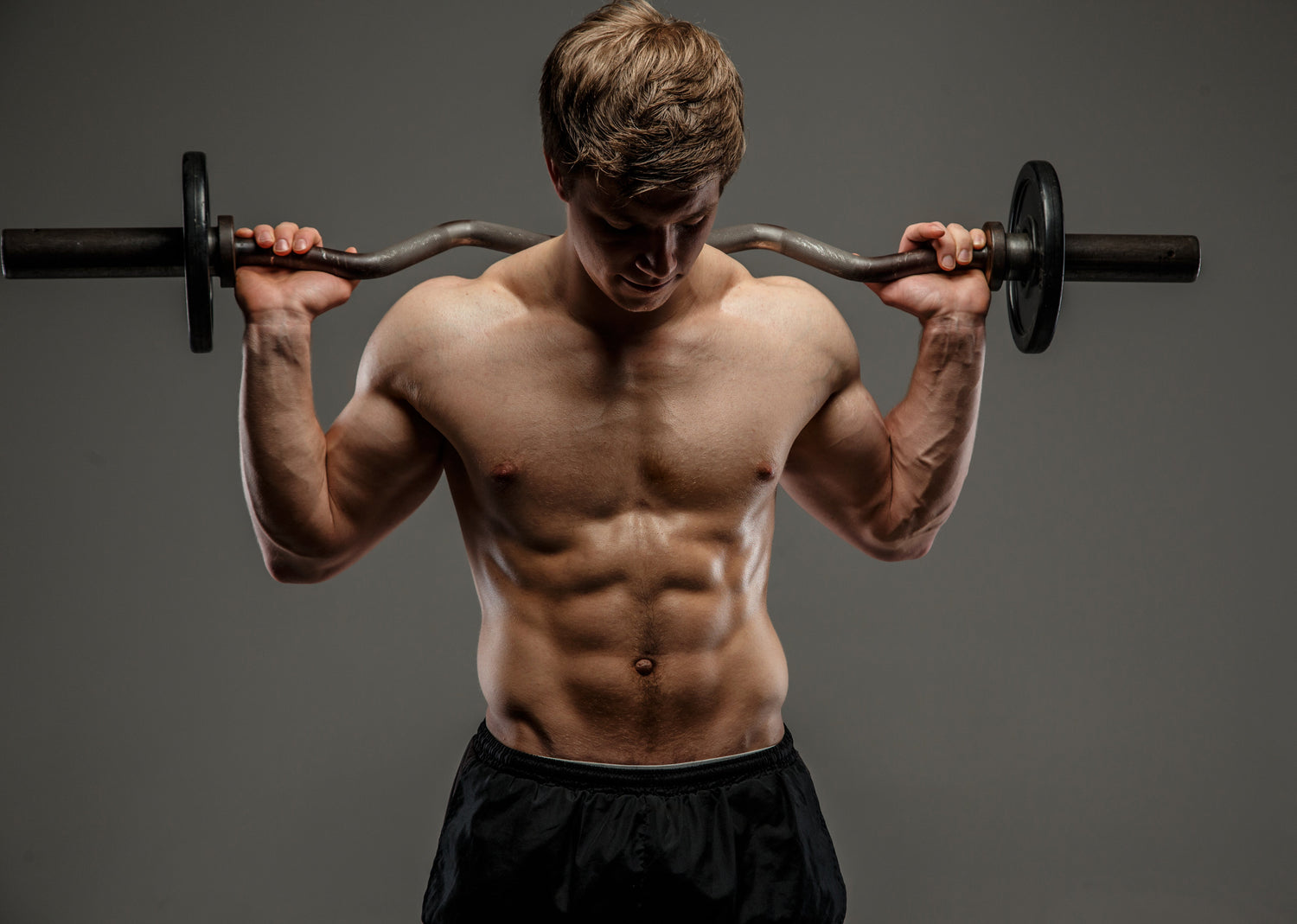 Shirtless man performing a barbell curl, highlighting muscular arms and defined core against a neutral studio background.