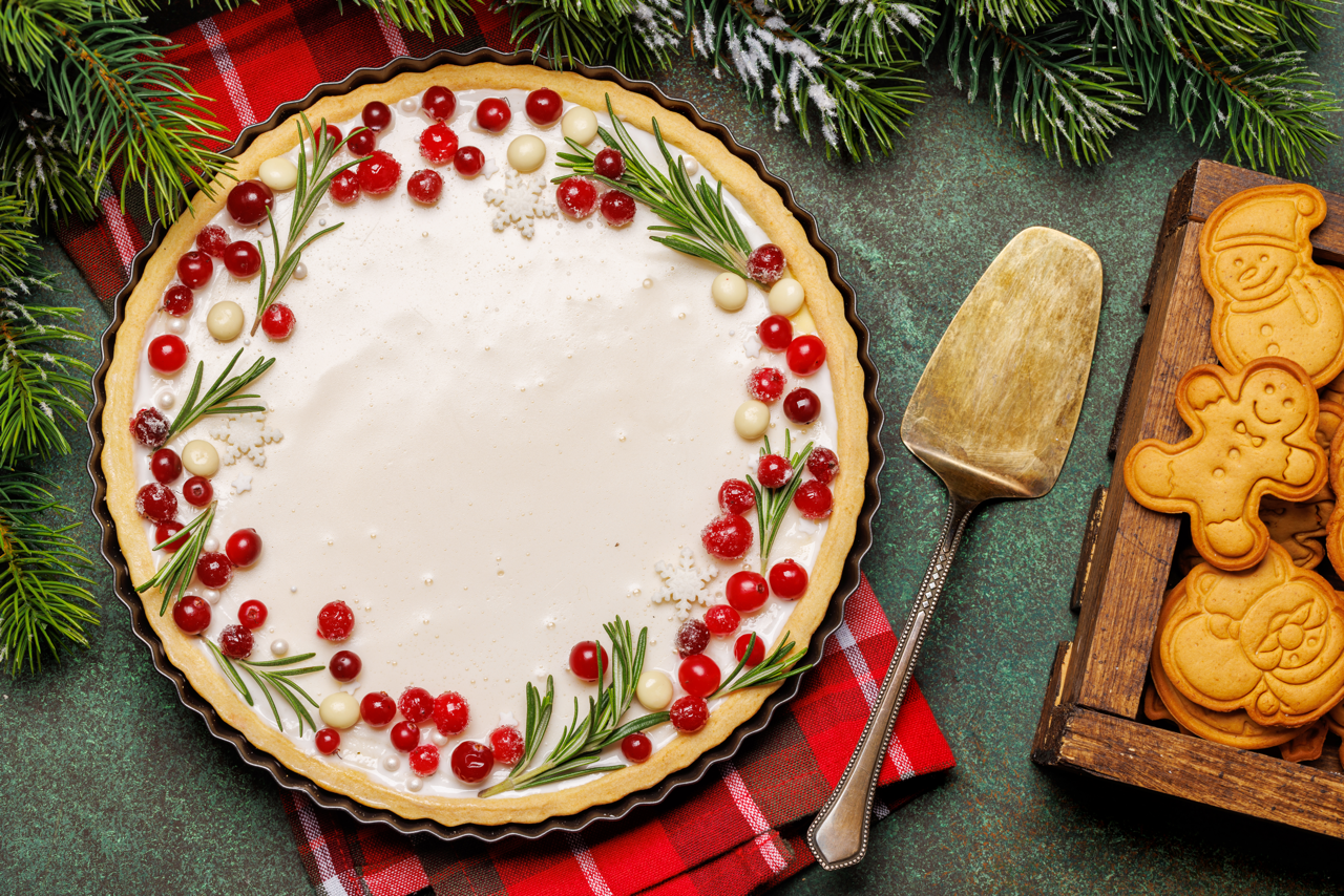 Gluten-Free Gingerbread Crust White Chocolate tart decorated with rosemary sprigs and red berries on a festive table, with gingerbread cookies and a cake server nearby.