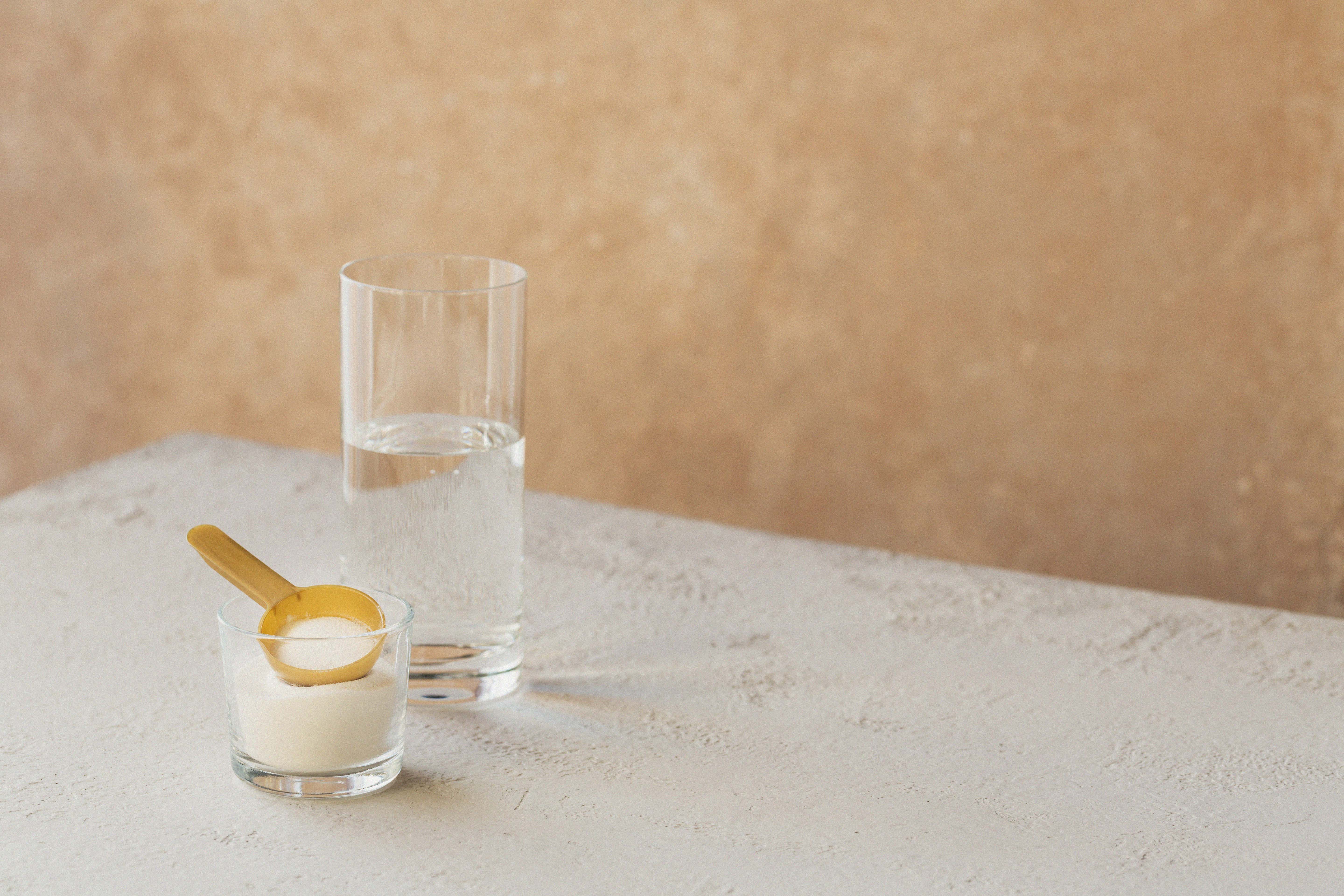 A small glass containing white powder with a gold measuring scoop beside a clear glass of water on a neutral textured surface.