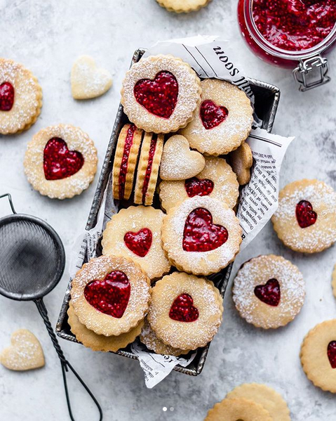 Heart-shaped jam sandwich cookies dusted with powdered sugar in a tray, with extra cookies, a sieve, and a jar of red jam on a light surface.