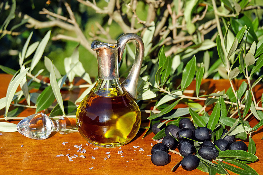 Glass bottle of olive oil with olives and leaves on a wooden surface, with coarse salt scattered nearby.