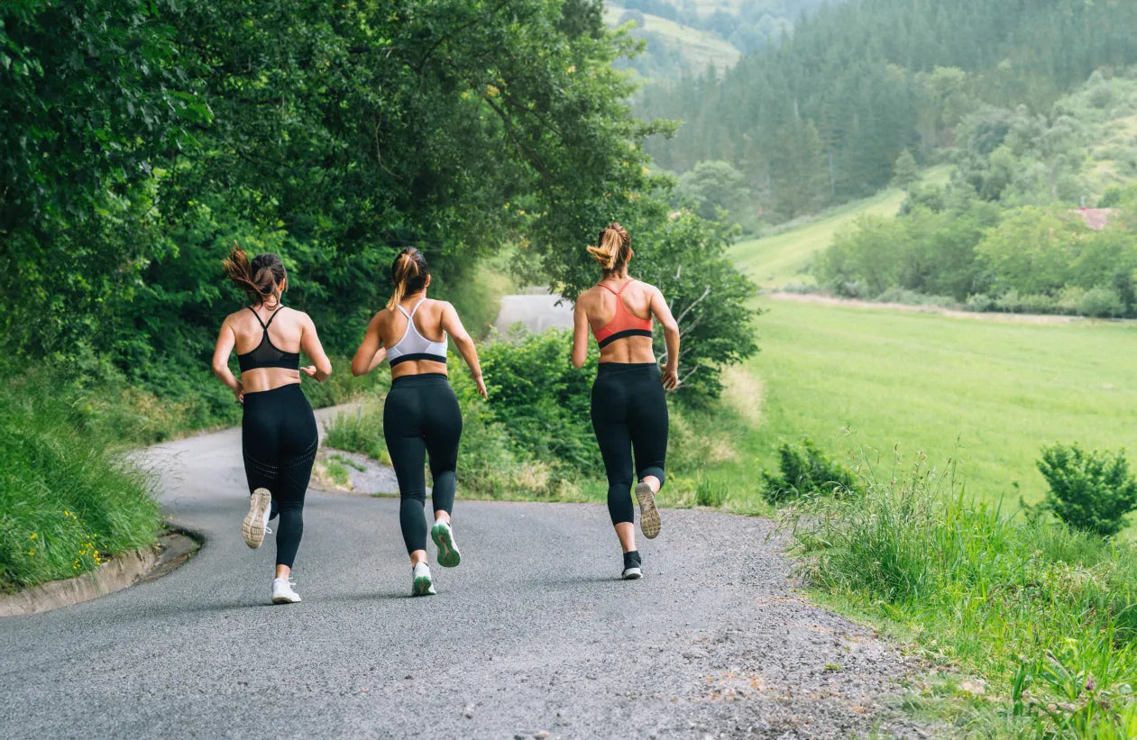 Three women in athletic wear run along a winding road surrounded by lush greenery and rolling hills