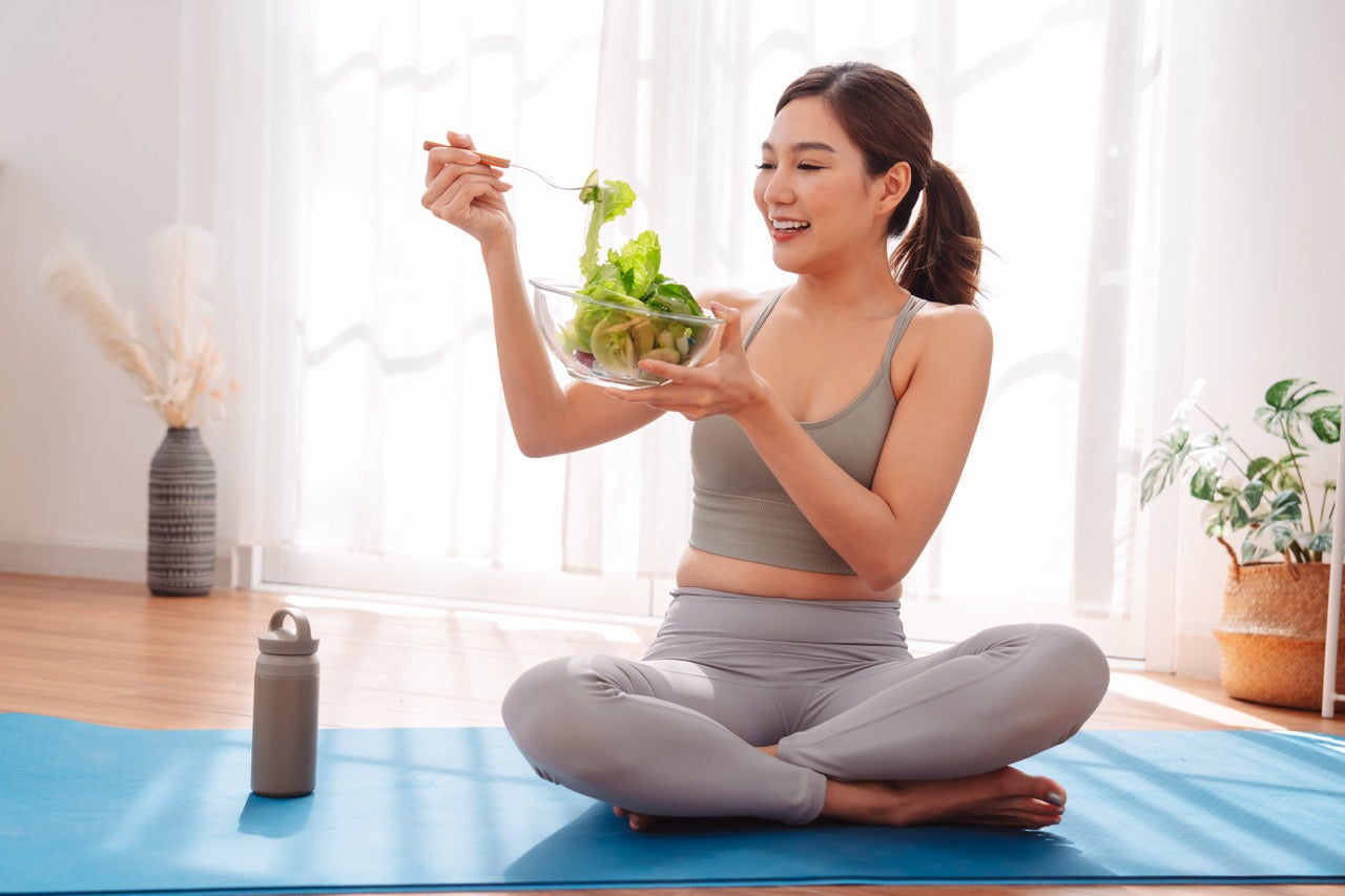 Woman sitting cross-legged on a yoga mat, smiling while eating a fresh salad, with a water bottle nearby in a bright, minimal indoor setting.
