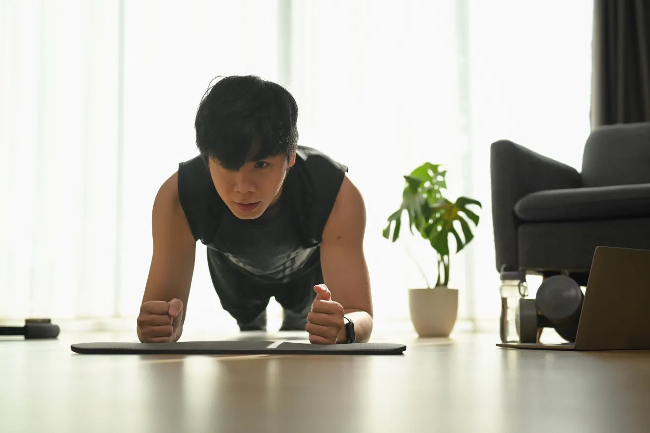 A young man in a sleeveless workout shirt holds a plank position on a yoga mat at home, with focused expression. A potted plant, dumbbells, water bottle, and laptop are visible in the background near a couch.