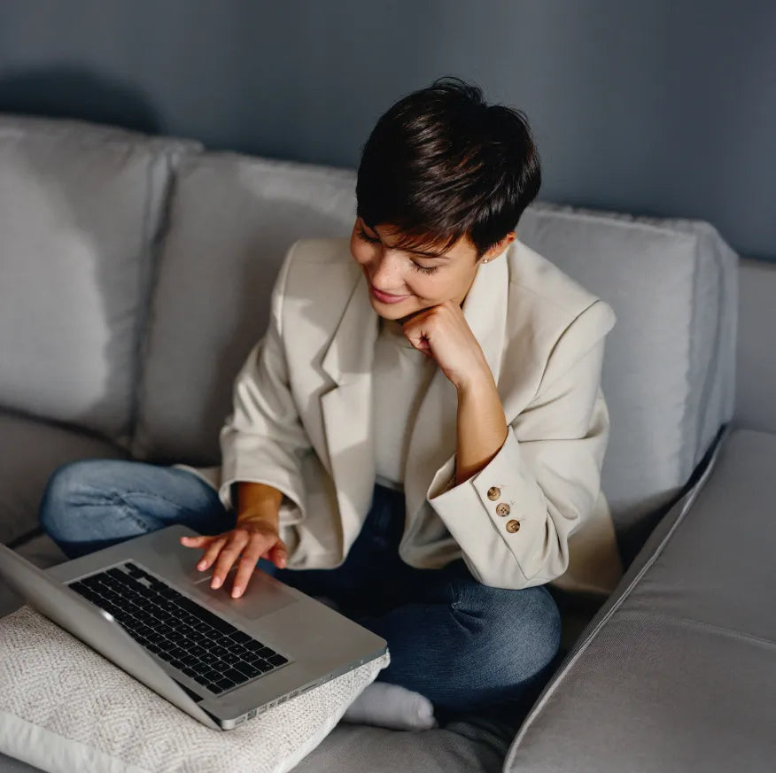 Smiling young woman sitting cross-legged on a couch, using a laptop placed on a pillow.