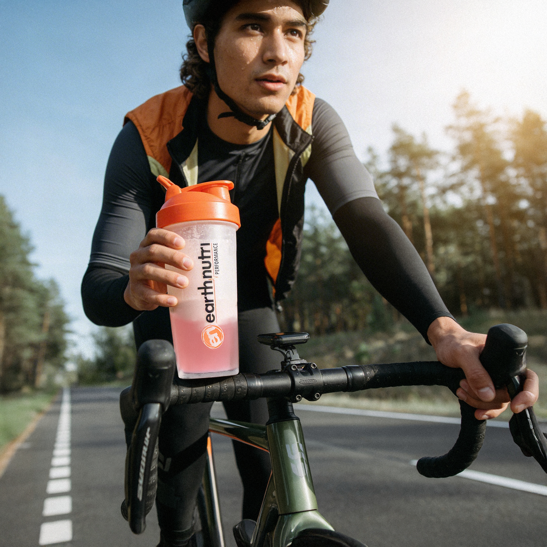 Person riding a bicycle holding an Earthnutshaker bottle on a road with trees in the background