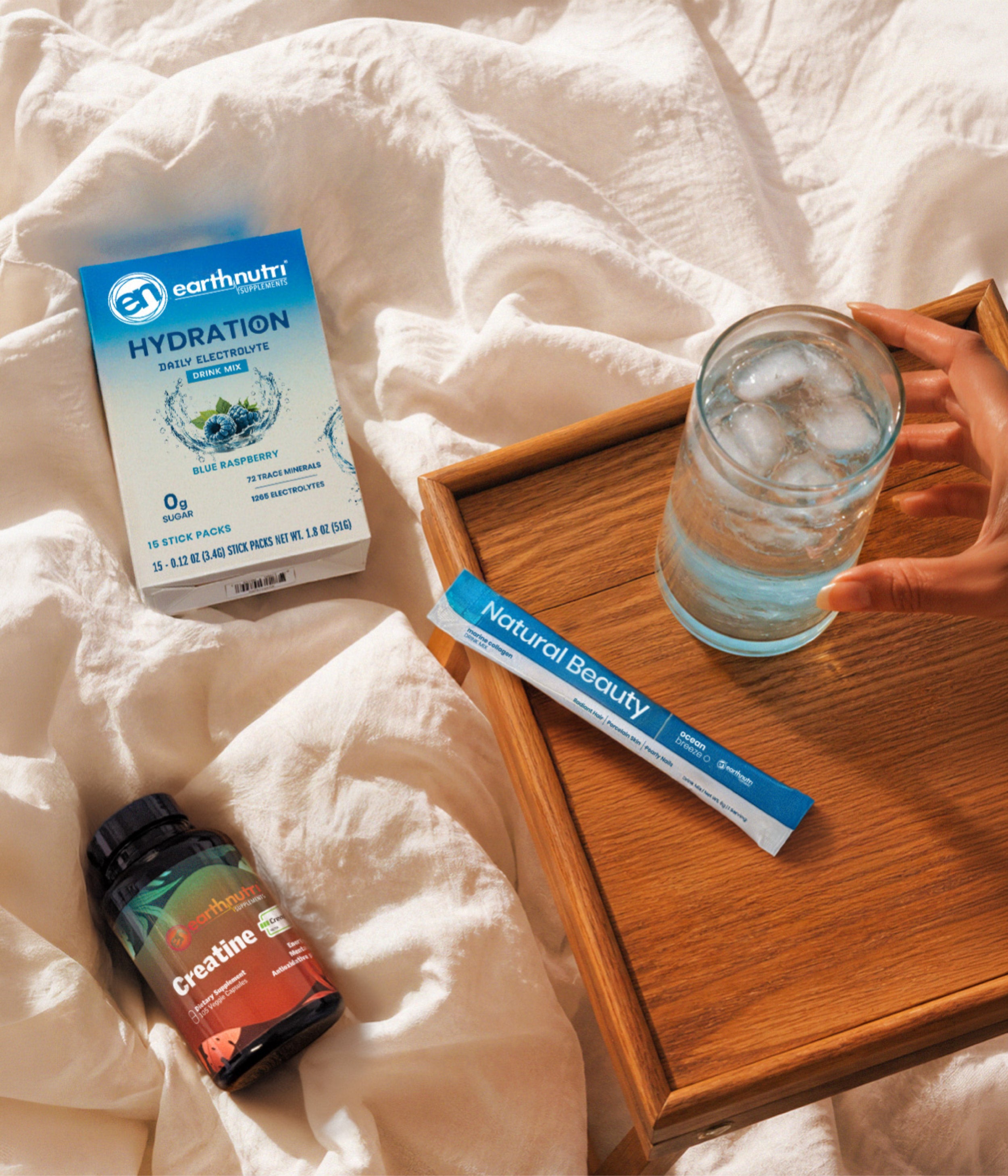 A styled flat-lay showing a box of Earthnutri Hydration electrolyte powder, a stick pack of Natural Beauty collagen powder, and a bottle of Creatine on white bed sheets next to a wooden tray with a glass of water being held by a hand.