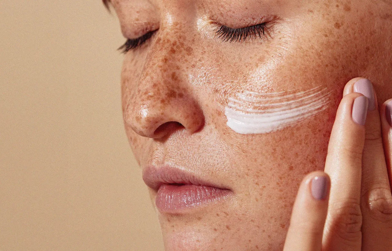 Close-up of a woman with freckles applying moisturizer to her cheek, highlighting natural skin texture and skincare routine