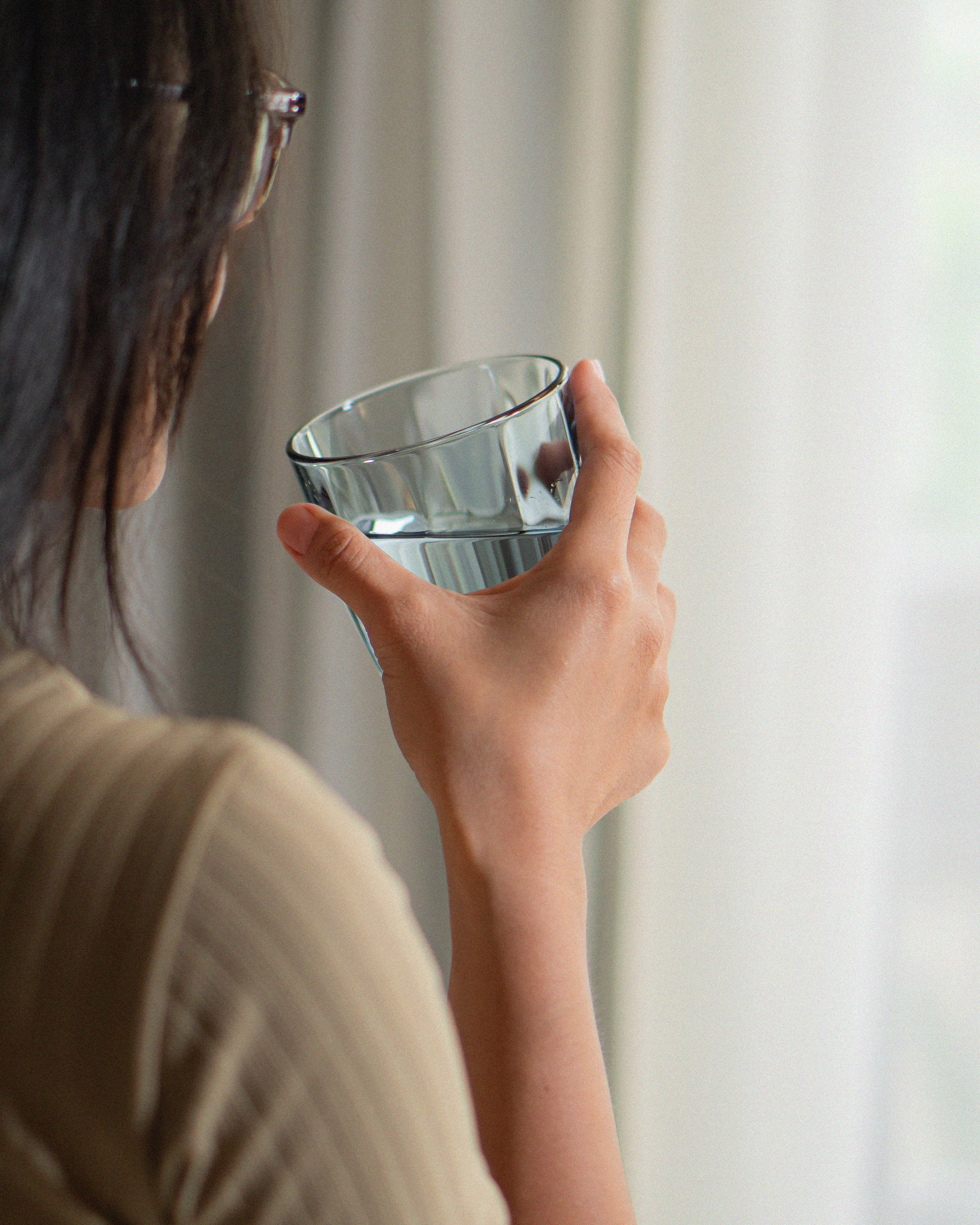 A person holding a glass of water near a window, viewed from behind.