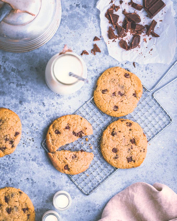 Chocolate chip cookies on a cooling rack with a glass bottle of milk, scattered chocolate pieces, and a soft cloth on a light surface.