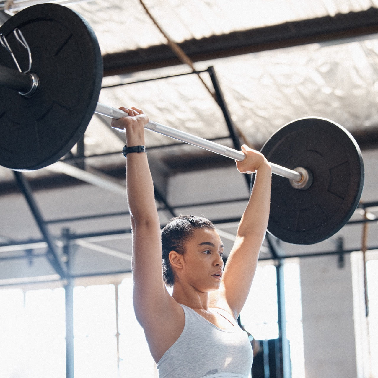Woman lifting a barbell overhead in a bright gym, demonstrating strength and controlled form.