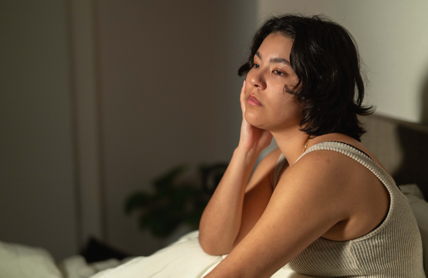 Person sitting on a bed in soft natural light rests their head on one hand, gazing thoughtfully to the side in a calm, intimate indoor setting.