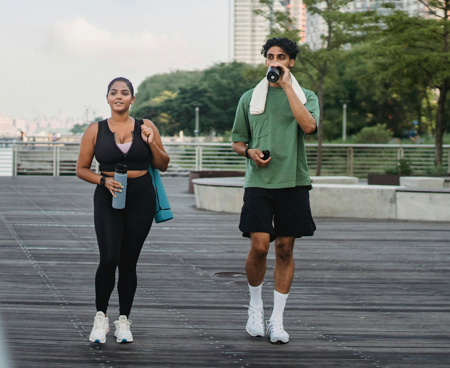 Two people walking on an outdoor boardwalk after exercise, each holding a water bottle. One drinks from a bottle while the other carries a yoga mat, with trees and city buildings in the background.