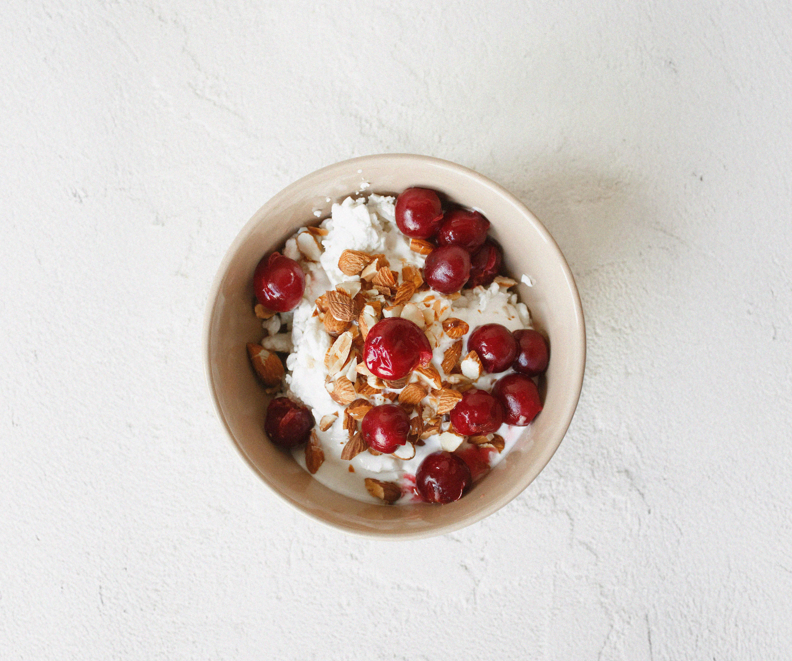 Top-down view of a ceramic bowl filled with creamy yogurt topped with red cherries and chopped almonds on a light textured background.