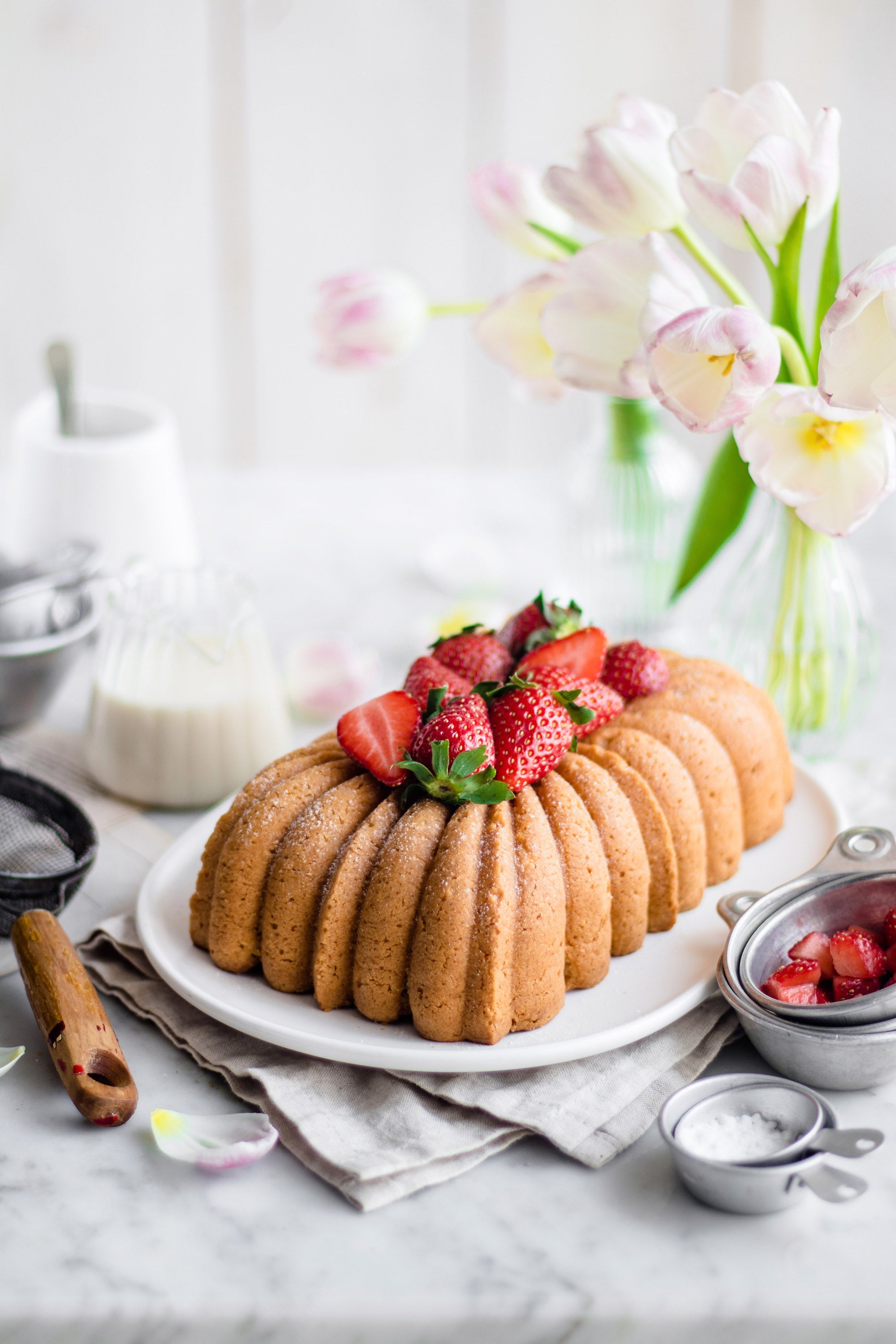 Bundt cake topped with fresh strawberries on a plate, with baking ingredients and flowers in the background.