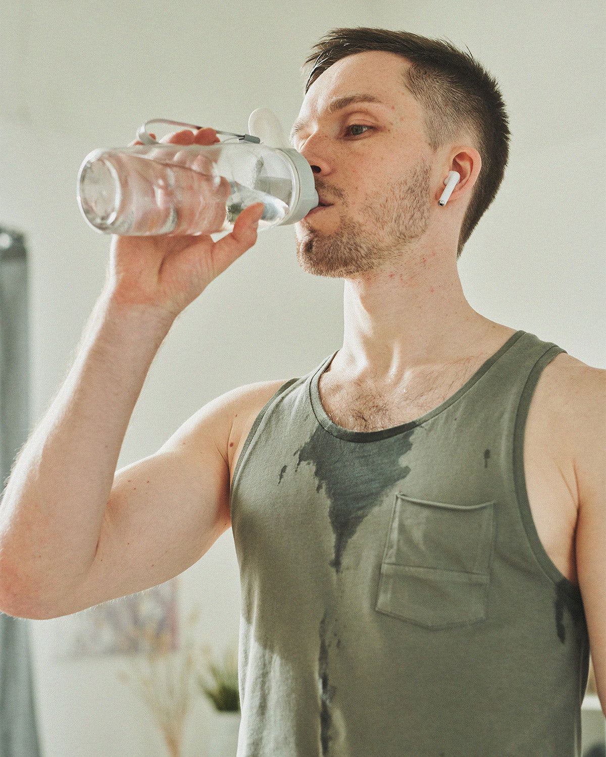 A person in a gray tank top drinks water from a clear reusable bottle after exercising. Sweat is visible on their shirt, and they wear a wireless earbud while standing indoors.