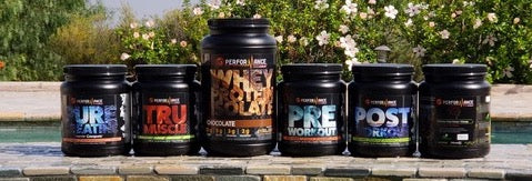 Lineup of supplement tubs displayed outdoors on a ledge, labeled products including hydration, muscle, pre-workout, and post-workout formulas with greenery in the background.
