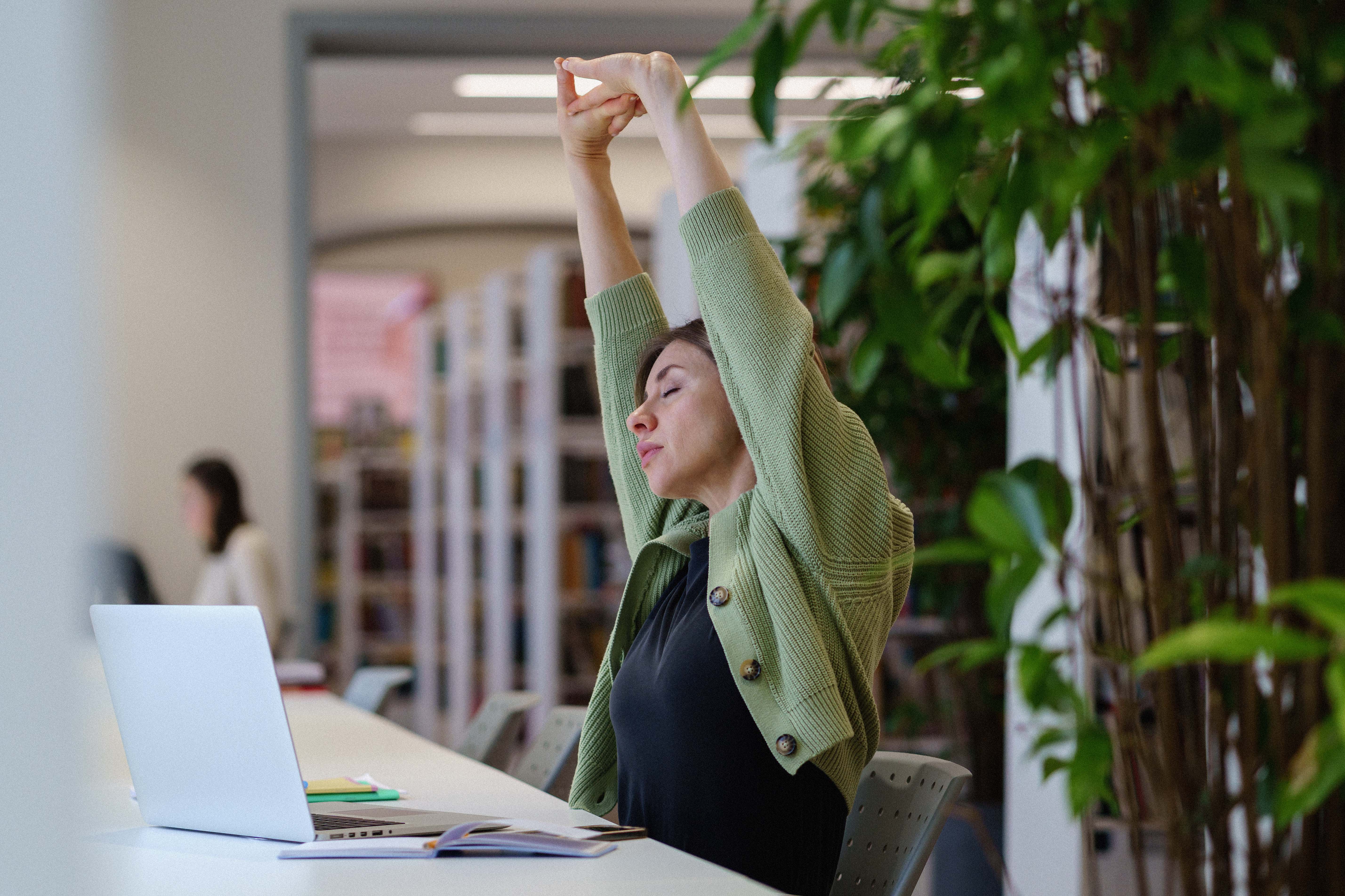 Woman seated at a desk in a bright workspace stretches her arms overhead beside an open laptop, eyes closed, with indoor plants in the background.