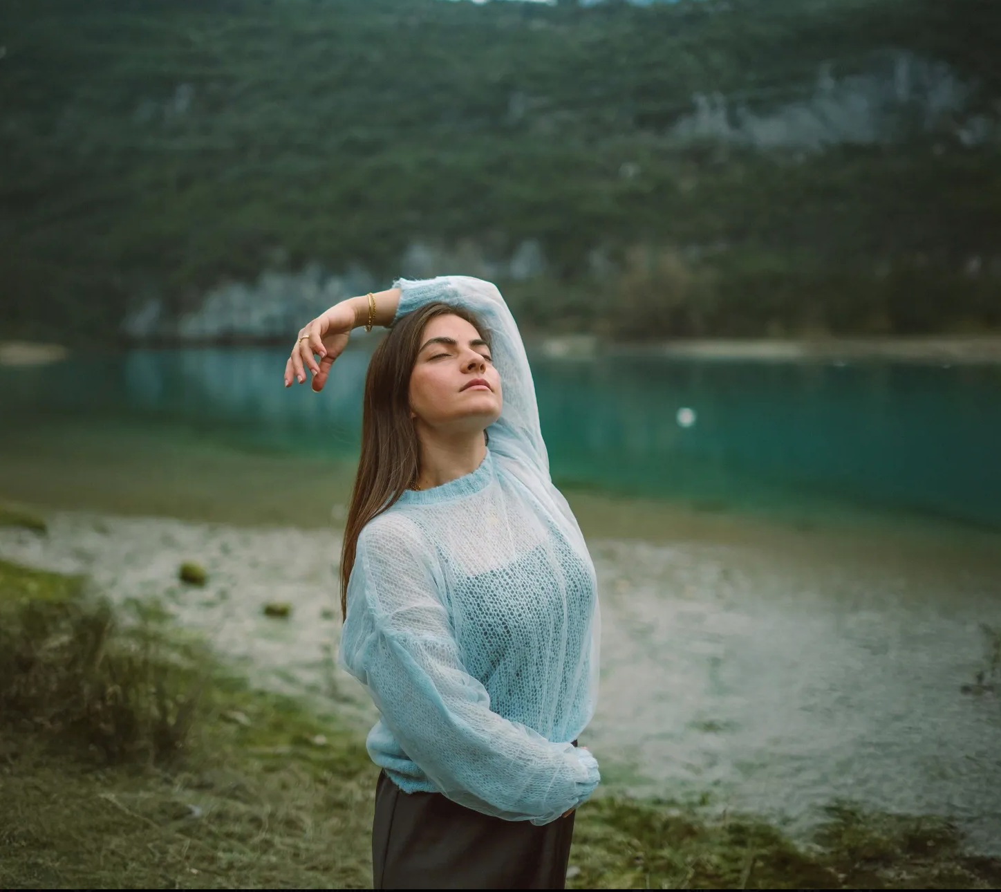 Woman in a sheer light-blue sweater stands by a calm turquoise lake with eyes closed and arm raised, appearing relaxed and mindful against a forested hillside backdrop.
