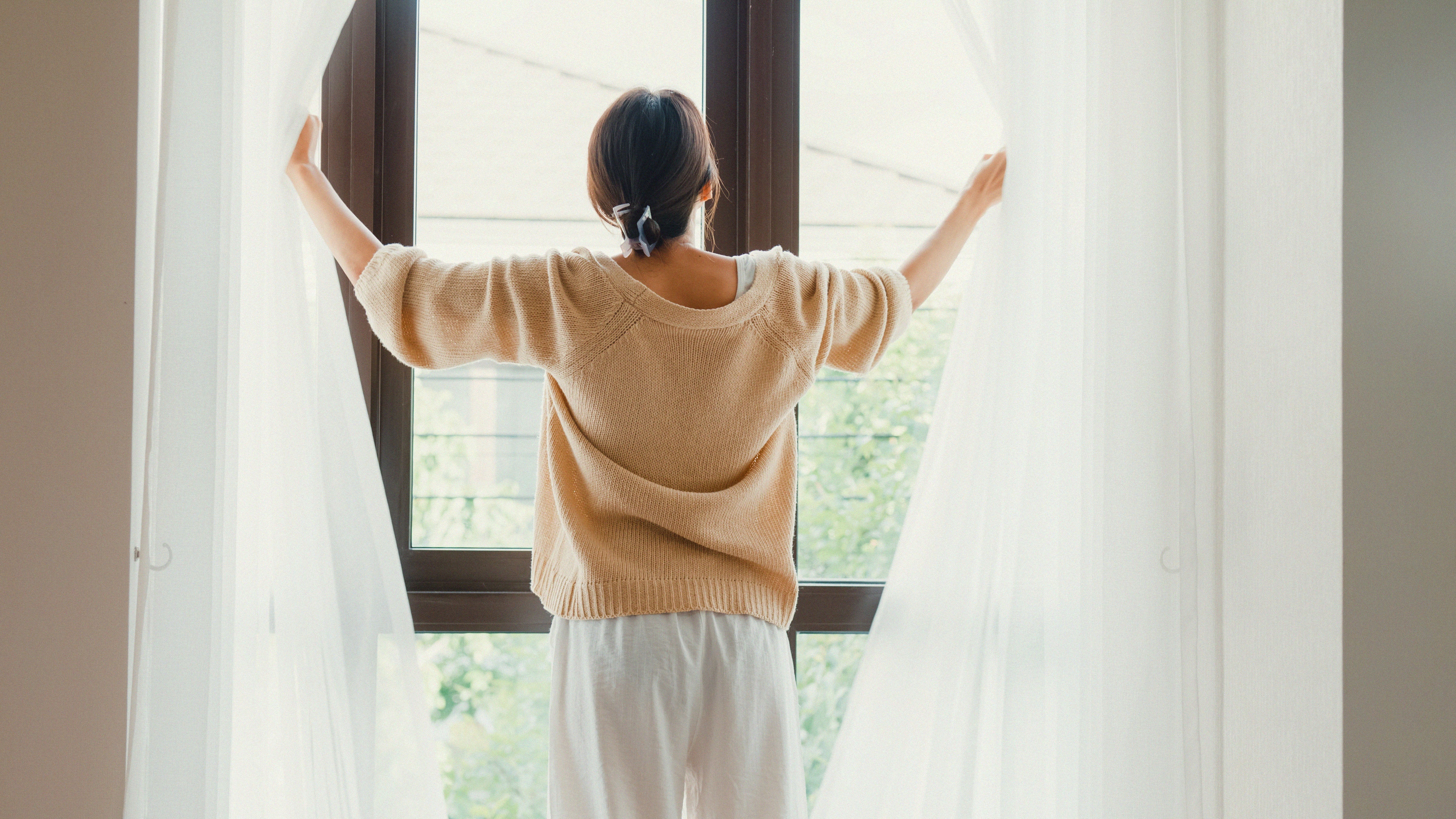 A woman stands in front of a large window, pulling open sheer white curtains to let in bright morning light, with greenery visible outside.