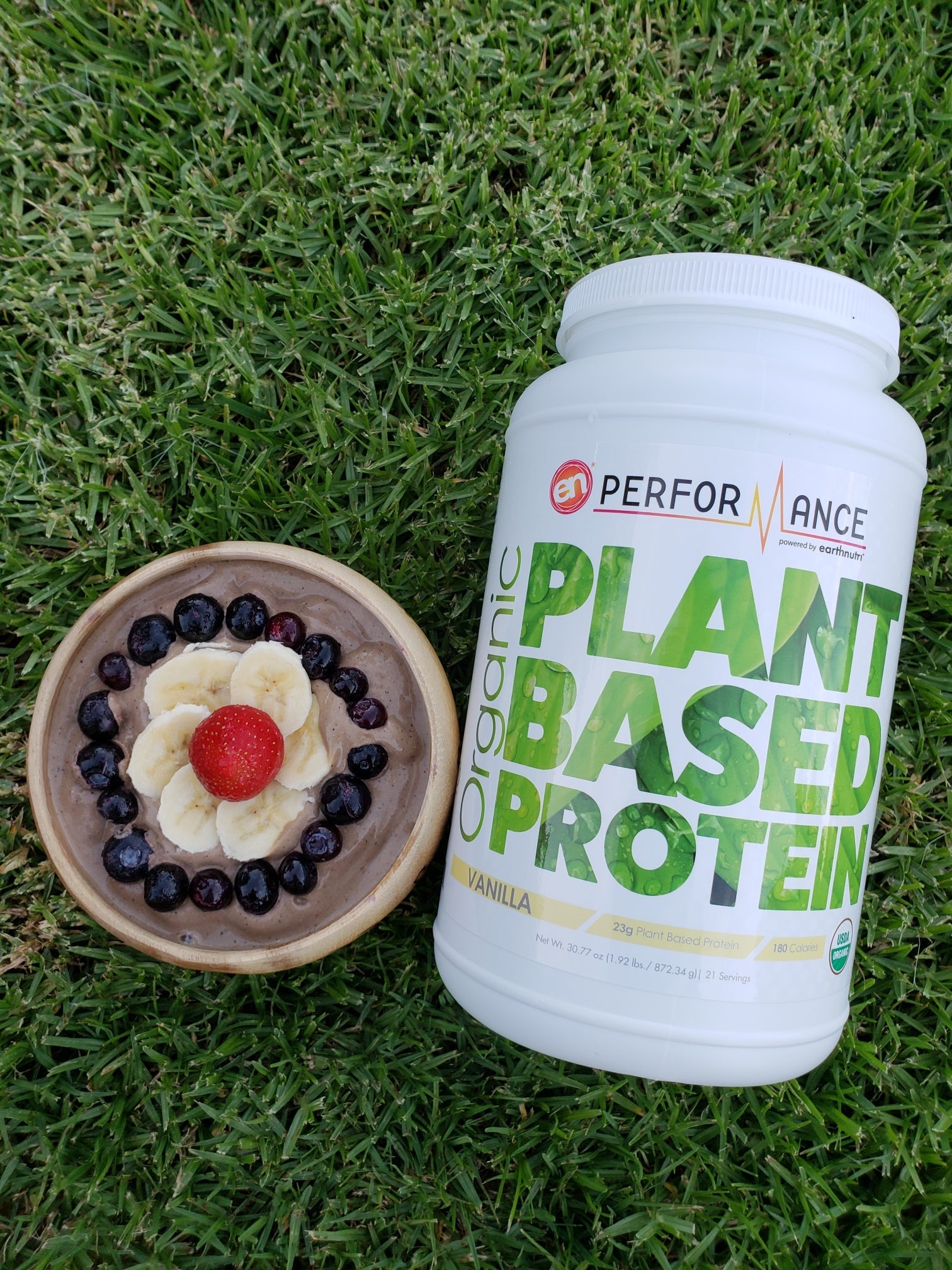 Container of organic plant-based protein on grass next to a bowl of oatmeal topped with banana slices, blueberries, and a strawberry.
