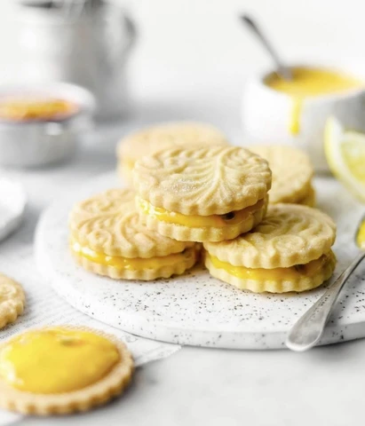 Stack of sandwich cookies filled with lemon cream on a plate, with lemon curd and utensils nearby. Gluten Free Lemon Passion Fruit Cookies