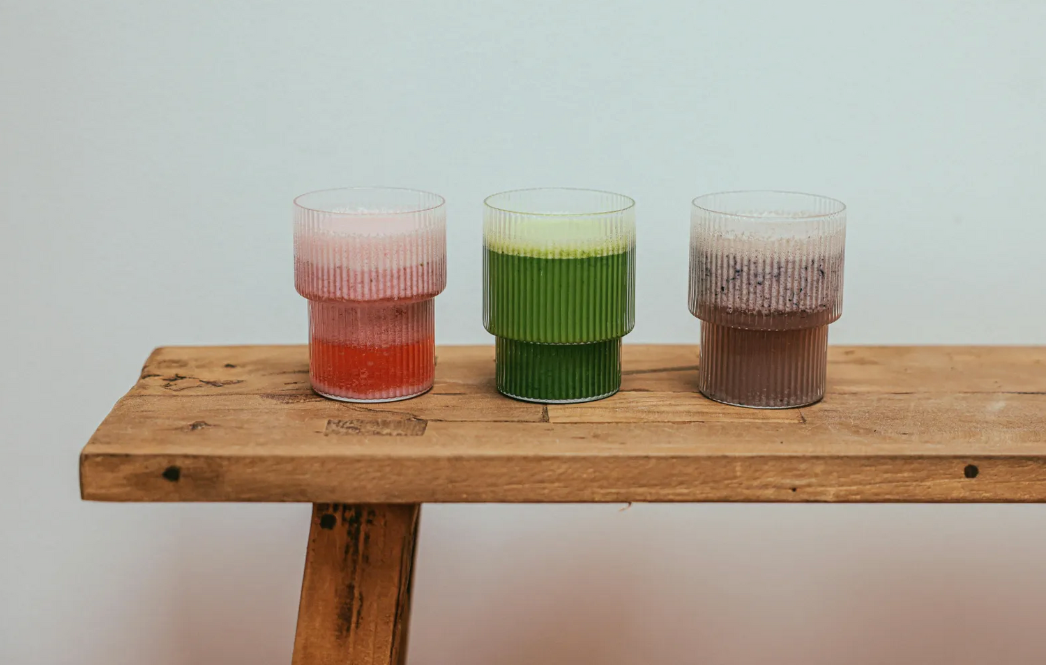 Three ribbed glass cups filled with layered smoothies—pink and red, green, and chocolate—sit side by side on a rustic wooden bench against a plain white wall.