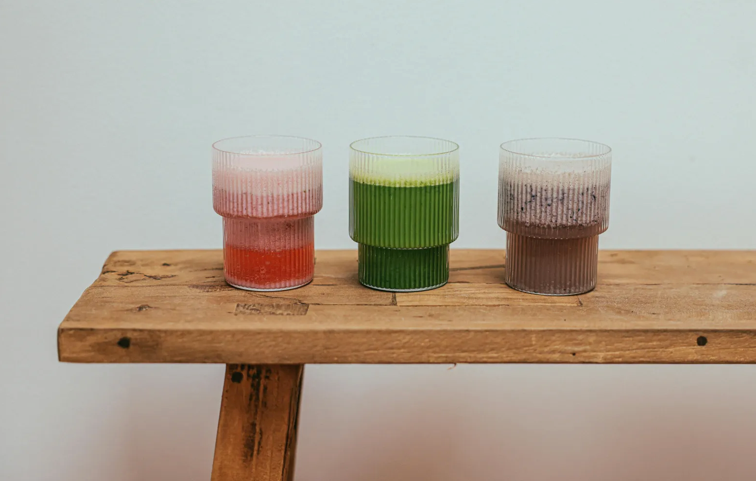 Three ribbed glass cups filled with layered smoothies—pink and red, green, and chocolate—sit side by side on a rustic wooden bench against a plain white wall.