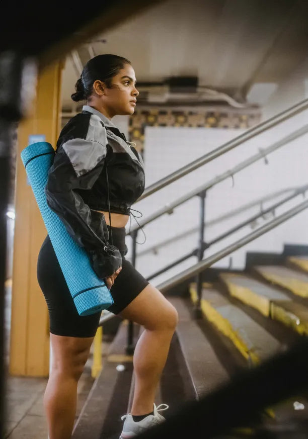 A woman in athletic wear carries a rolled-up blue yoga mat while walking up subway stairs, looking focused and determined.