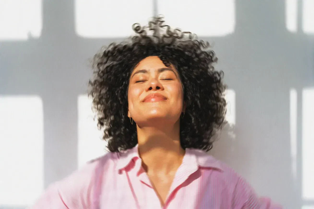 A joyful woman with curly hair stands in sunlight with her eyes closed, smiling peacefully, symbolizing natural energy and well-being without caffeine.