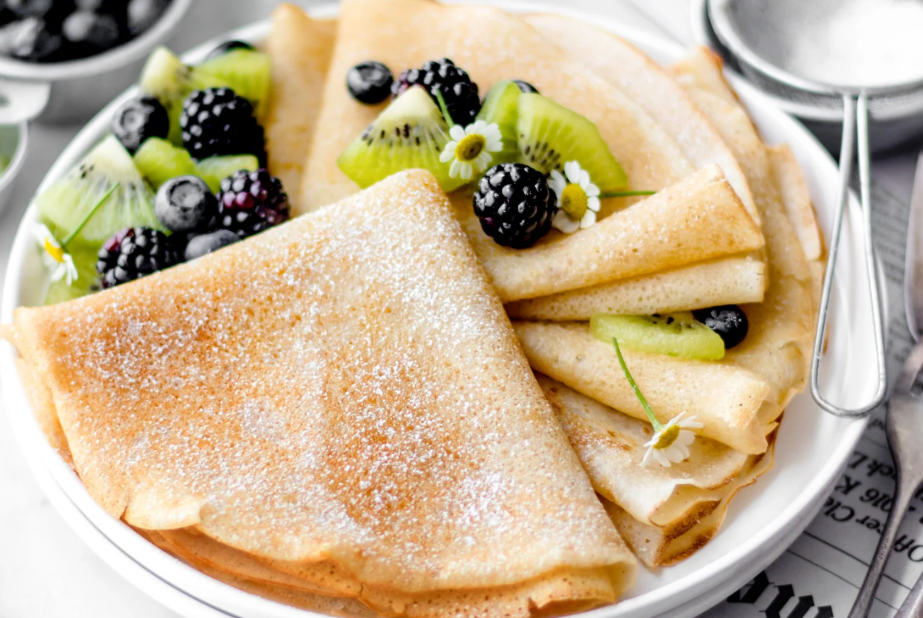 Thin crepes folded and dusted with powdered sugar on a plate, served with fresh kiwi slices, blackberries, and blueberries on a light tabletop. Gluten-Free French Crepes