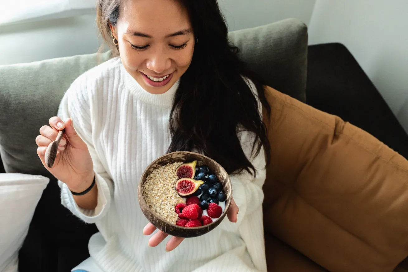 A smiling woman in a white sweater holds a wooden bowl of oatmeal topped with figs, blueberries, and raspberries while sitting on a couch.