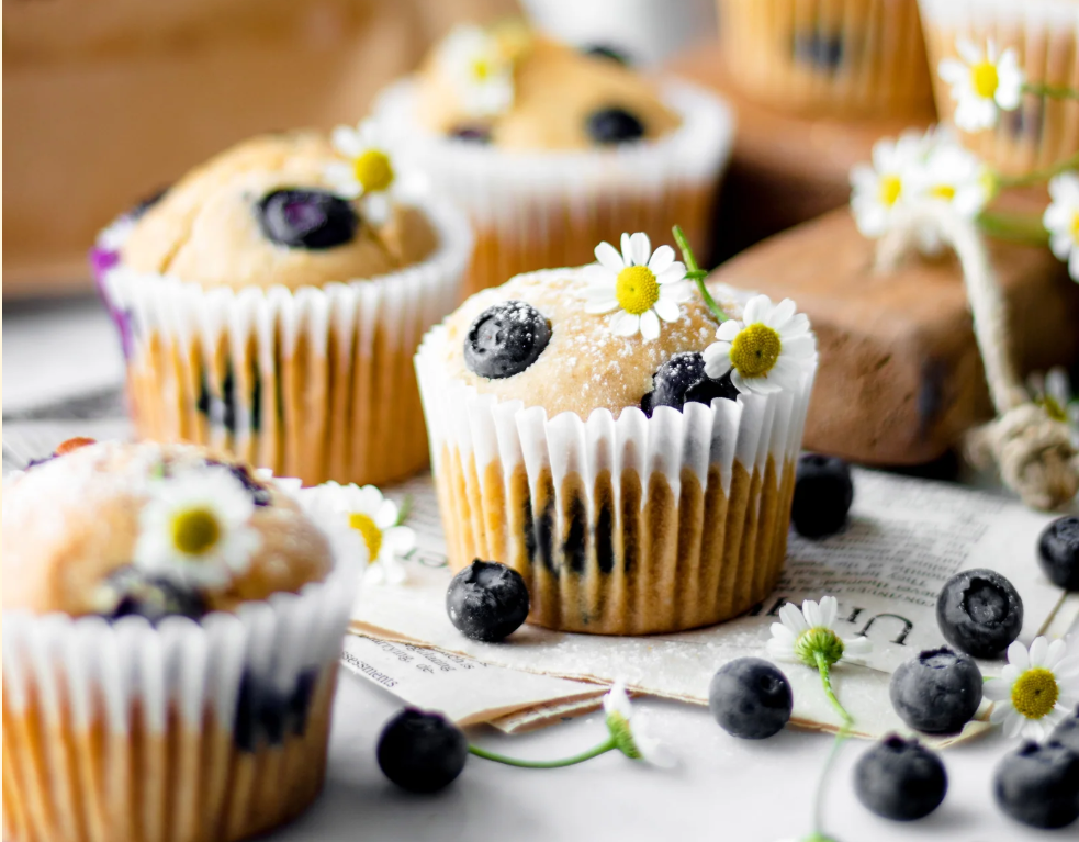 Blueberry muffins in white liners dusted with powdered sugar, topped with small white flowers, with scattered blueberries on a light surface.. Gluten-Free Blueberry Muffins