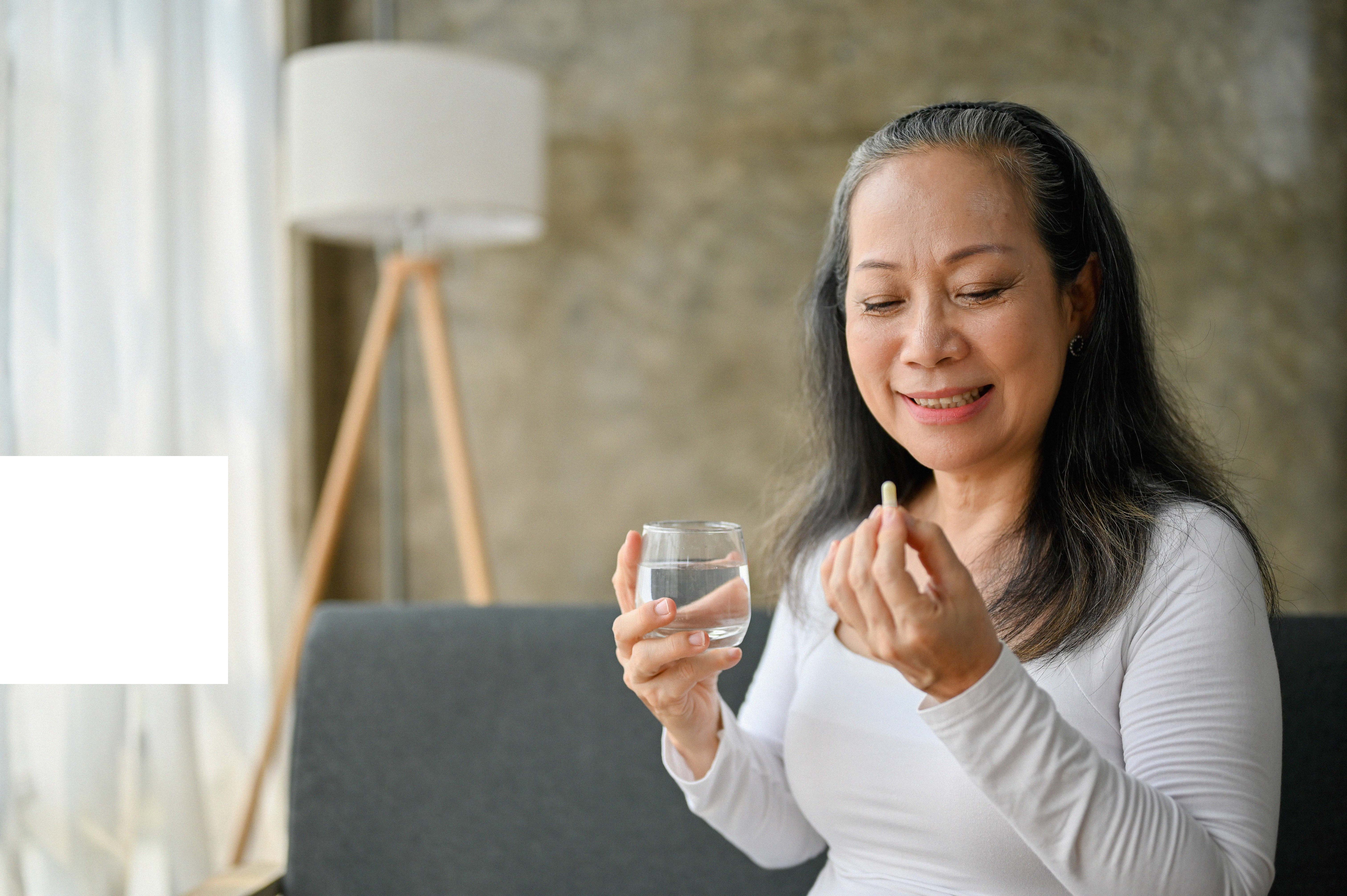 A smiling middle-aged woman sitting on a couch holds a glass of water in one hand and a capsule in the other, preparing to take a supplement in a softly lit living room.