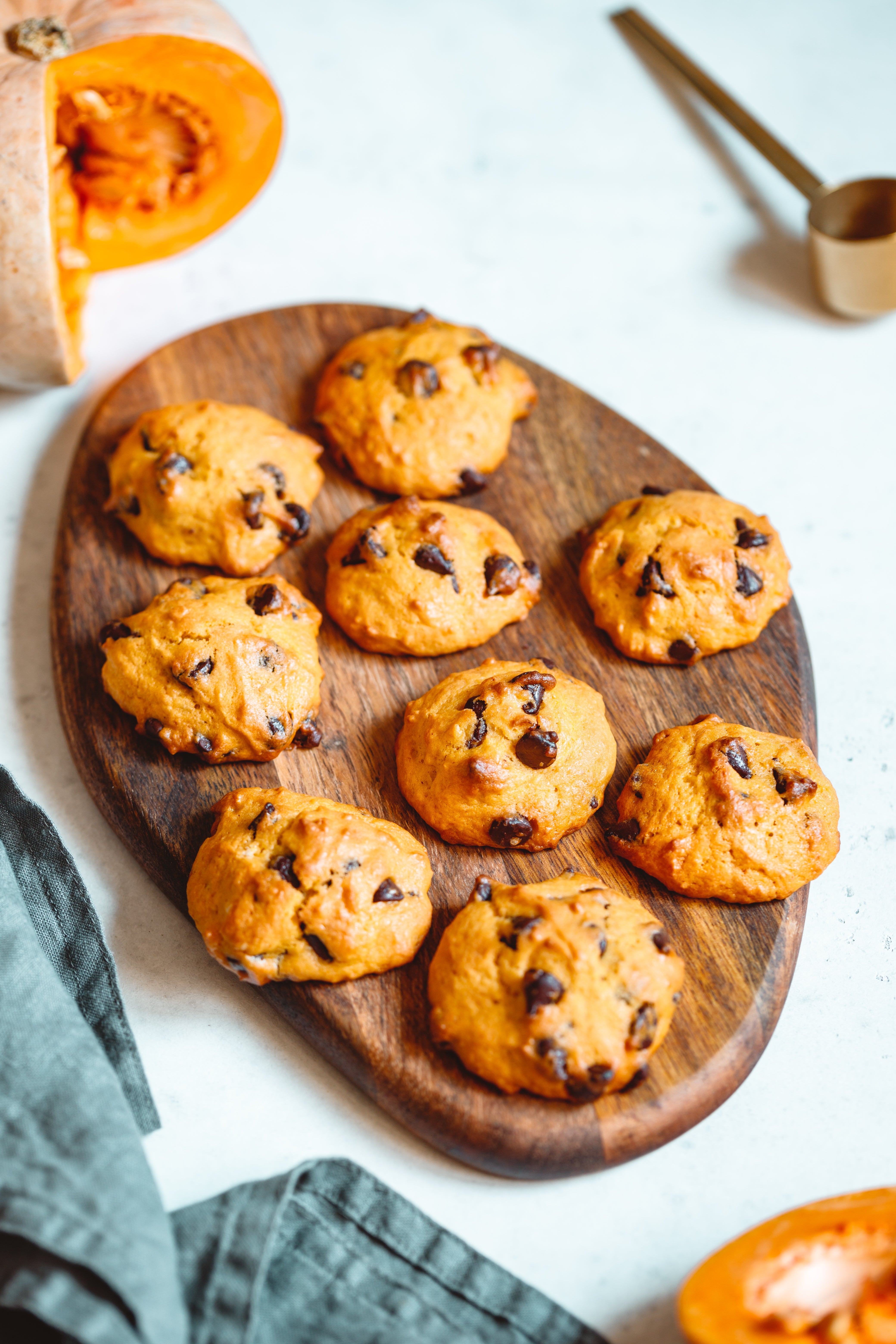 Freshly baked gluten-free pumpkin chocolate chip cookies arranged on a wooden serving board, with a halved pumpkin and measuring spoon in the background.