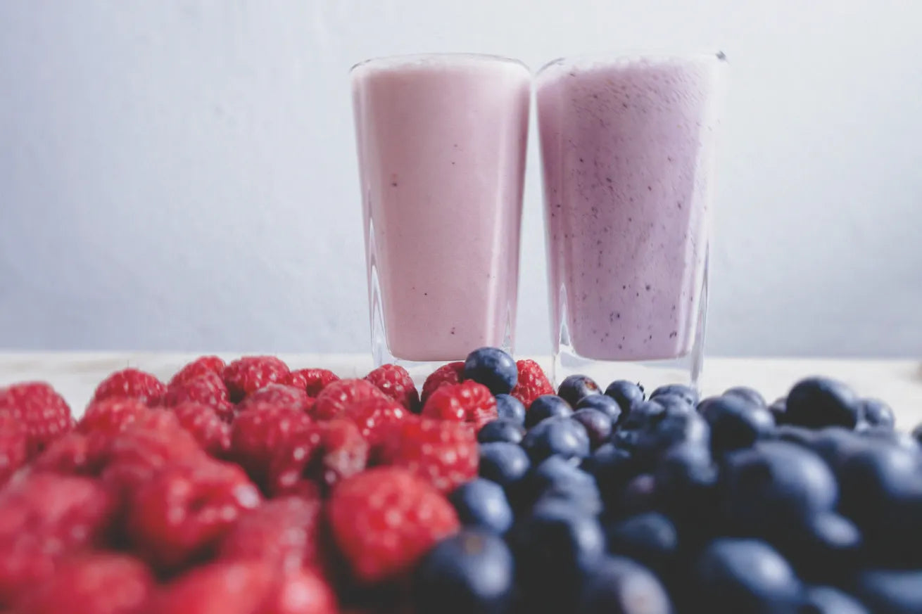 Two glasses of berry smoothies, one pink and one purple, with fresh raspberries and blueberries in the foreground.